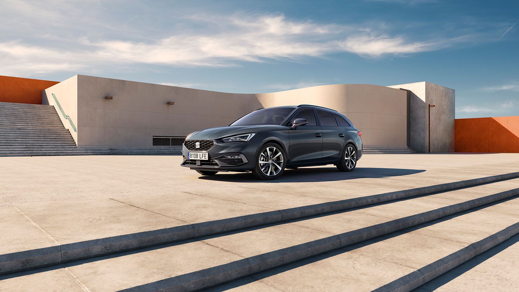 hree quarter view of grey 2024 SEAT Leon with matrix led headlamp lights, parked in front of modern architecture with orange and white walls and palm trees in the background